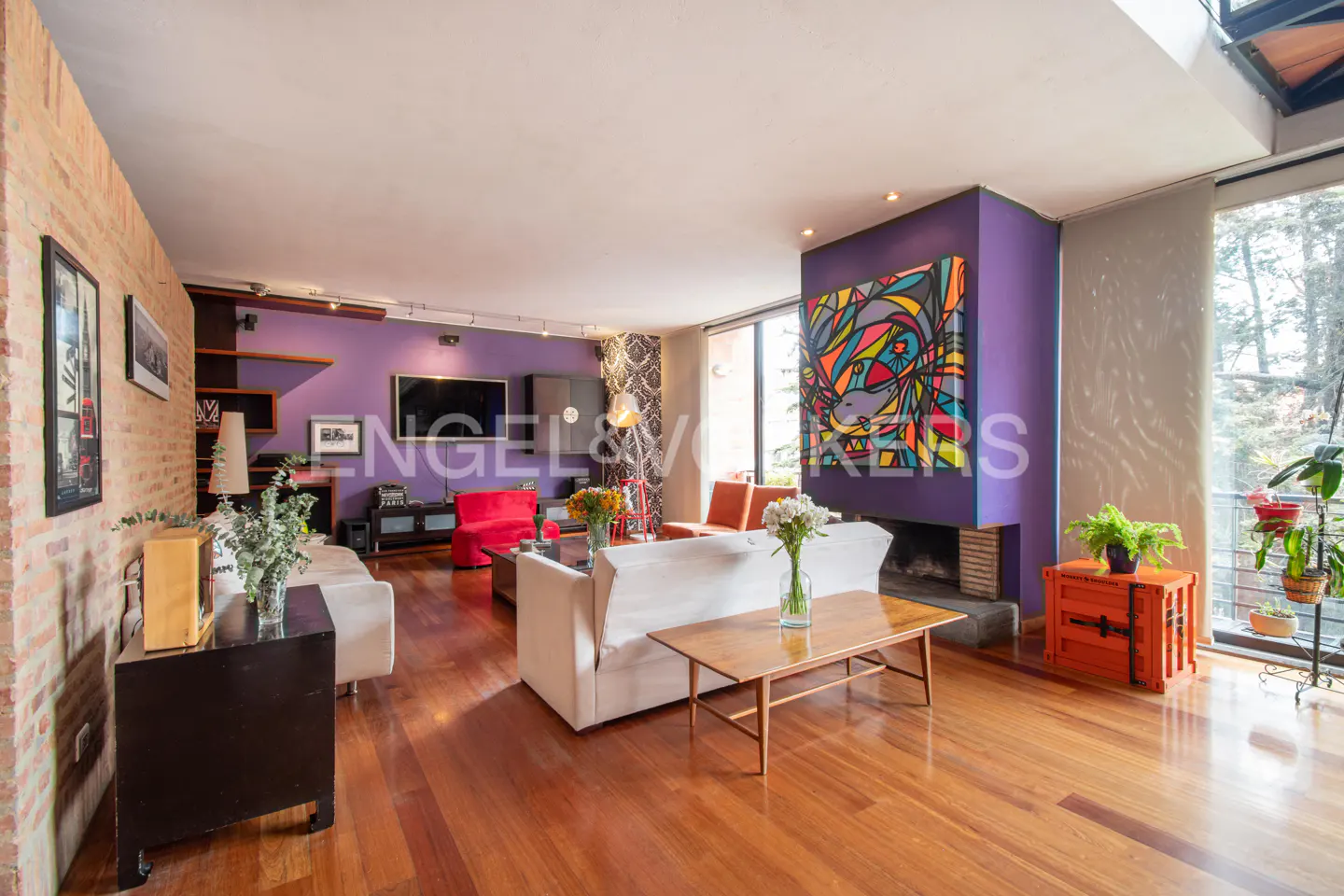 A spacious living room with hardwood floors, white sofas, a purple accent wall, and a colorful painting above the fireplace.