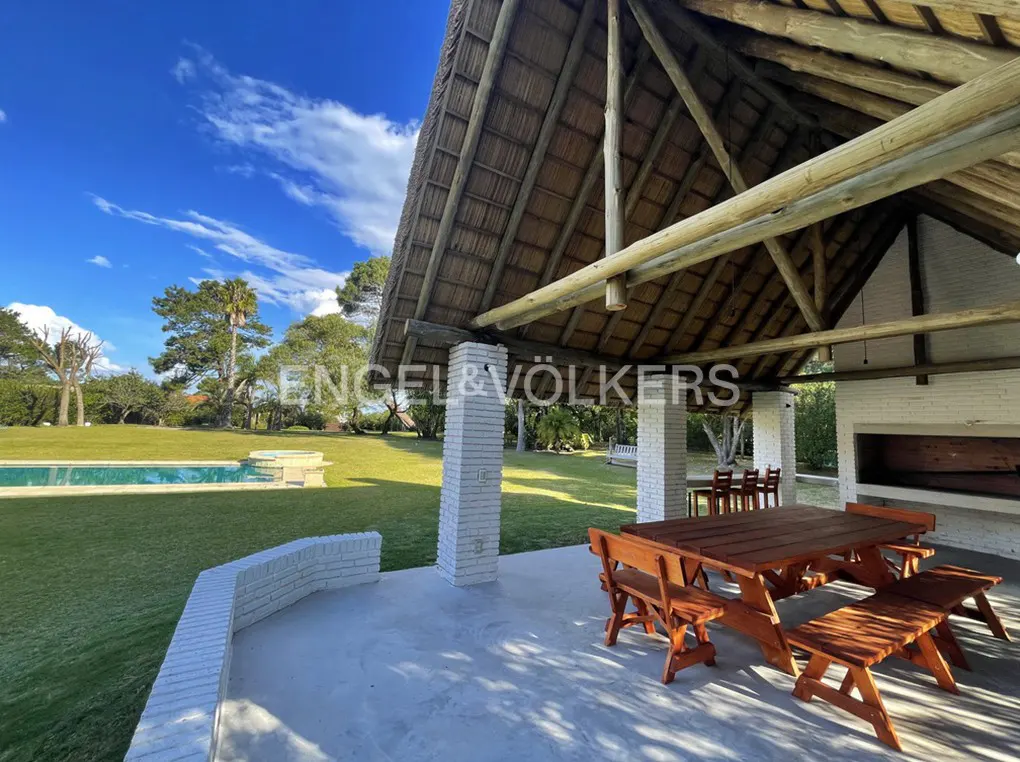 Outdoor dining area with a thatched roof, wooden table and benches, white brick pillars, and a view of a green lawn and pool.