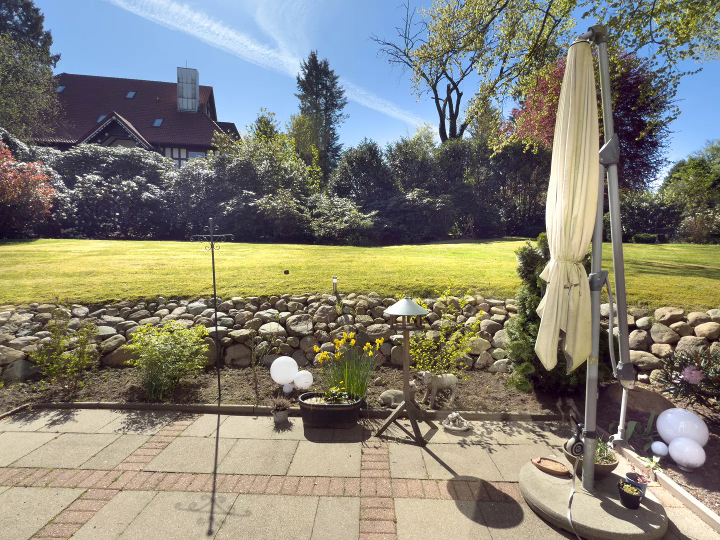 A backyard patio with a stone wall, green lawn, trees, and a house in the background. A closed beige umbrella stands on the patio.