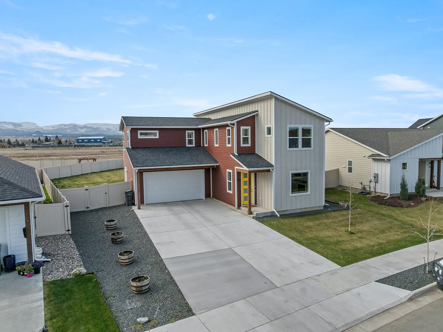 Two-story house with a yellow door, gray siding, and a maroon section. A long driveway leads to a white garage door. Blue sky.