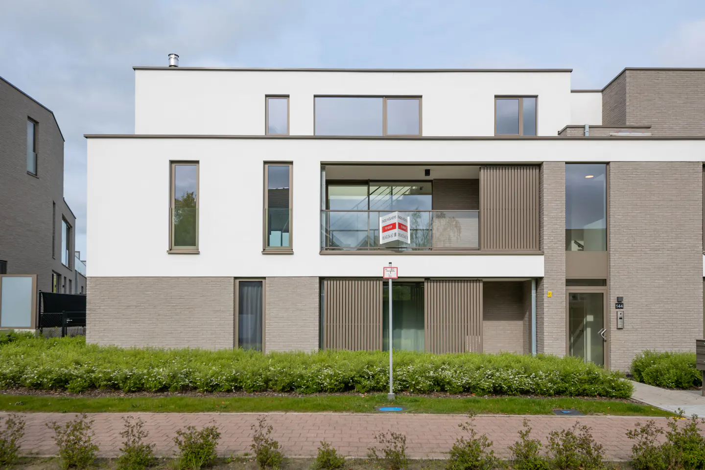 Modern townhouse with white and gray brick exterior. A "For Sale" sign hangs on the balcony. Green bushes line the front.