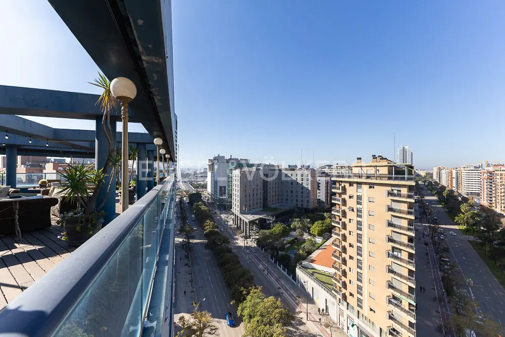View from a high-rise balcony with glass railings, overlooking a city street with buildings, trees, and cars under a clear blue sky.