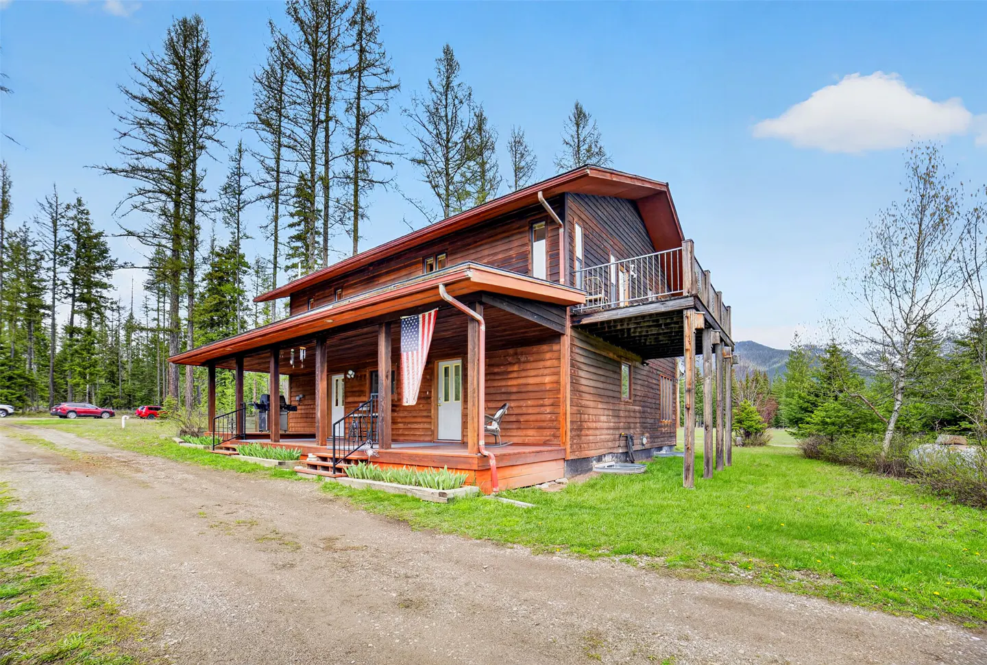 Two-story log cabin with a porch and balcony, an American flag, and a gravel driveway. Tall trees and a blue sky are in the background.