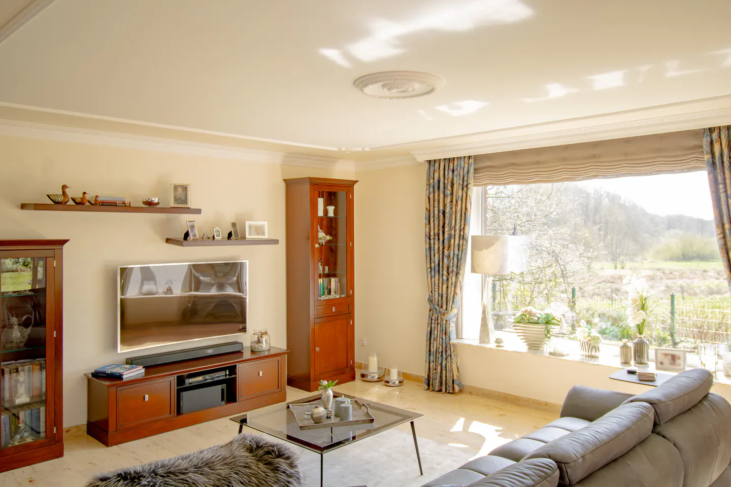 Living room with wood floors, brown cabinets, and a gray sofa. Large window with floral curtains overlooks a green landscape.