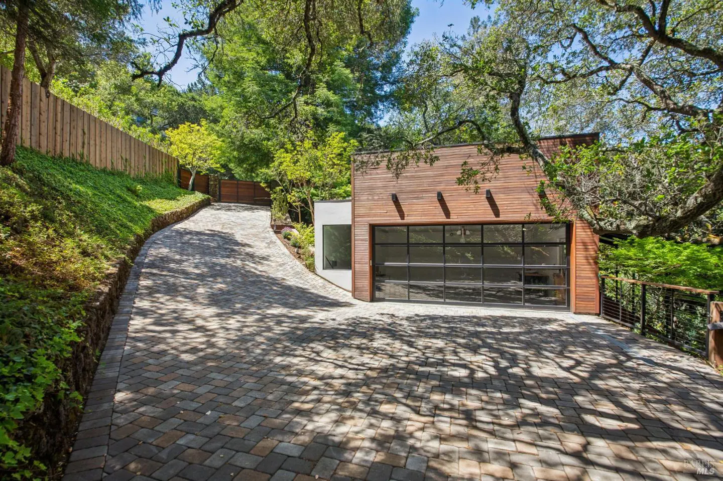 Exterior view of a modern house with a wood facade and a glass garage door, surrounded by trees and a brick driveway.