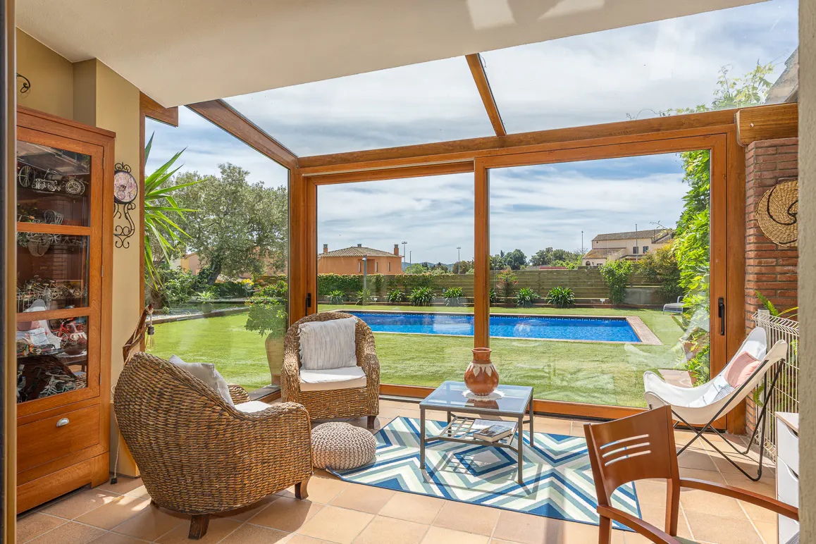 Sunroom with wicker chairs, glass table, and blue rug. Outside, a pool is surrounded by green grass and trees.