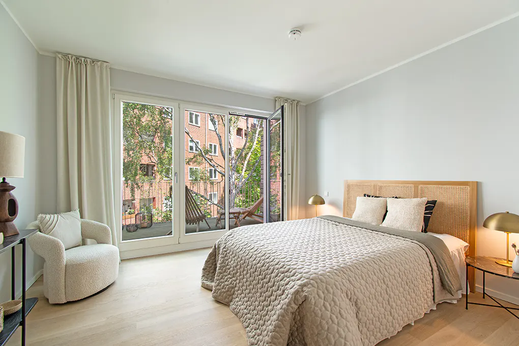 Bedroom with a bed, chair, and balcony. The bed has a wicker headboard and a beige quilt. The balcony has chairs and a view of trees and a brick building.