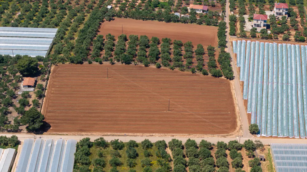 Aerial view of farmland with brown soil, green trees, greenhouses, and a few houses.