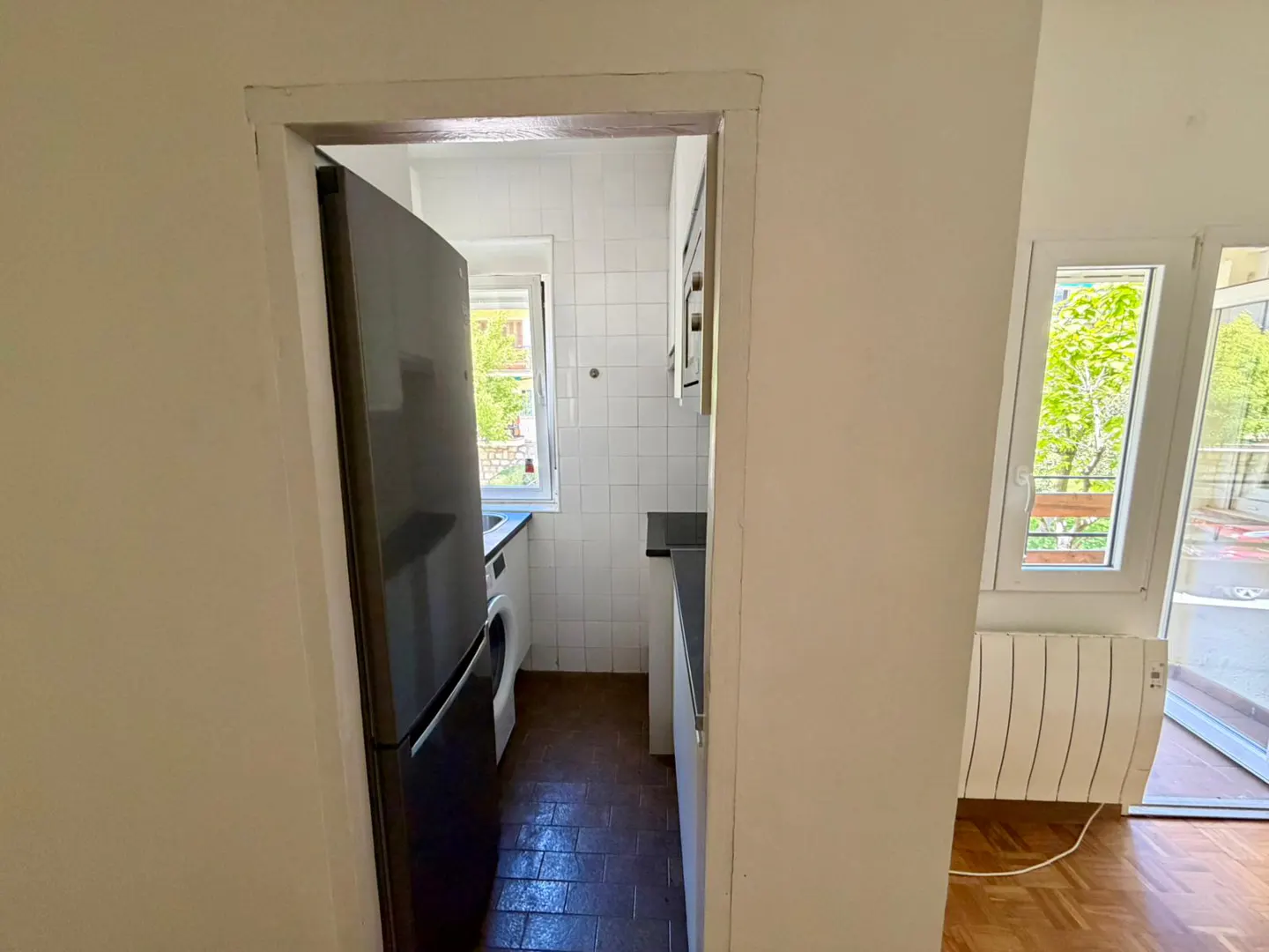 A view through a doorway into a small kitchen with a black refrigerator, white cabinets, and a window. A window and radiator are visible in the adjacent room.