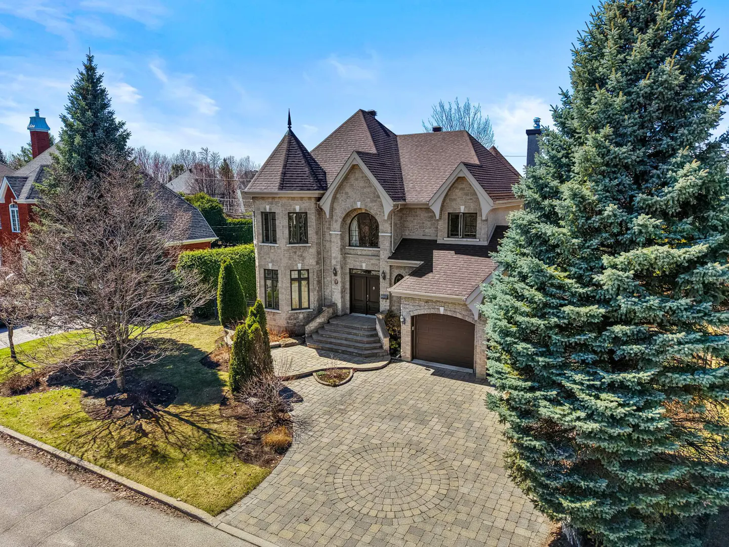 Aerial view of a large, two-story stone house with a brown roof and a circular driveway. Tall evergreen trees frame the house.
