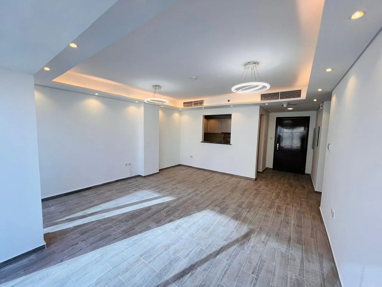 Spacious, empty living room with white walls, wood-look tile floors, modern circular pendant lights, and a kitchen pass-thru.