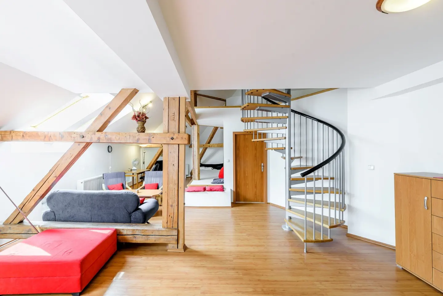 Modern loft interior with wooden beams, a grey sofa, a red ottoman, and a metal spiral staircase on a hardwood floor.