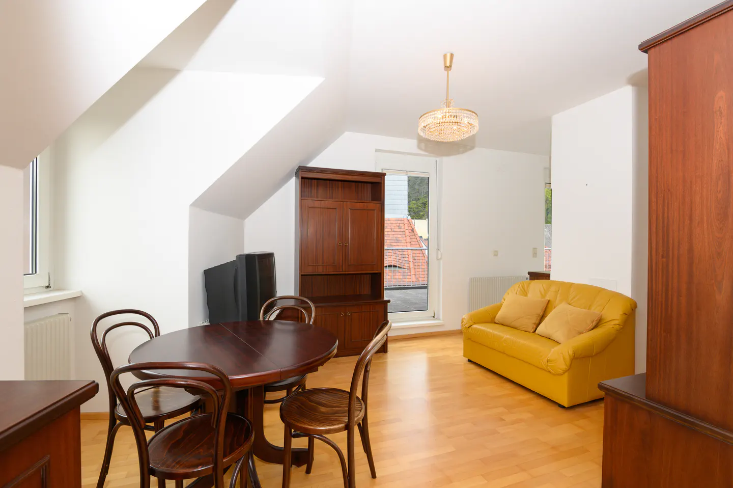 Attic apartment with wood floors, white walls, and a chandelier. A yellow sofa sits near a window, and a dark wood table and chairs are in the foreground.