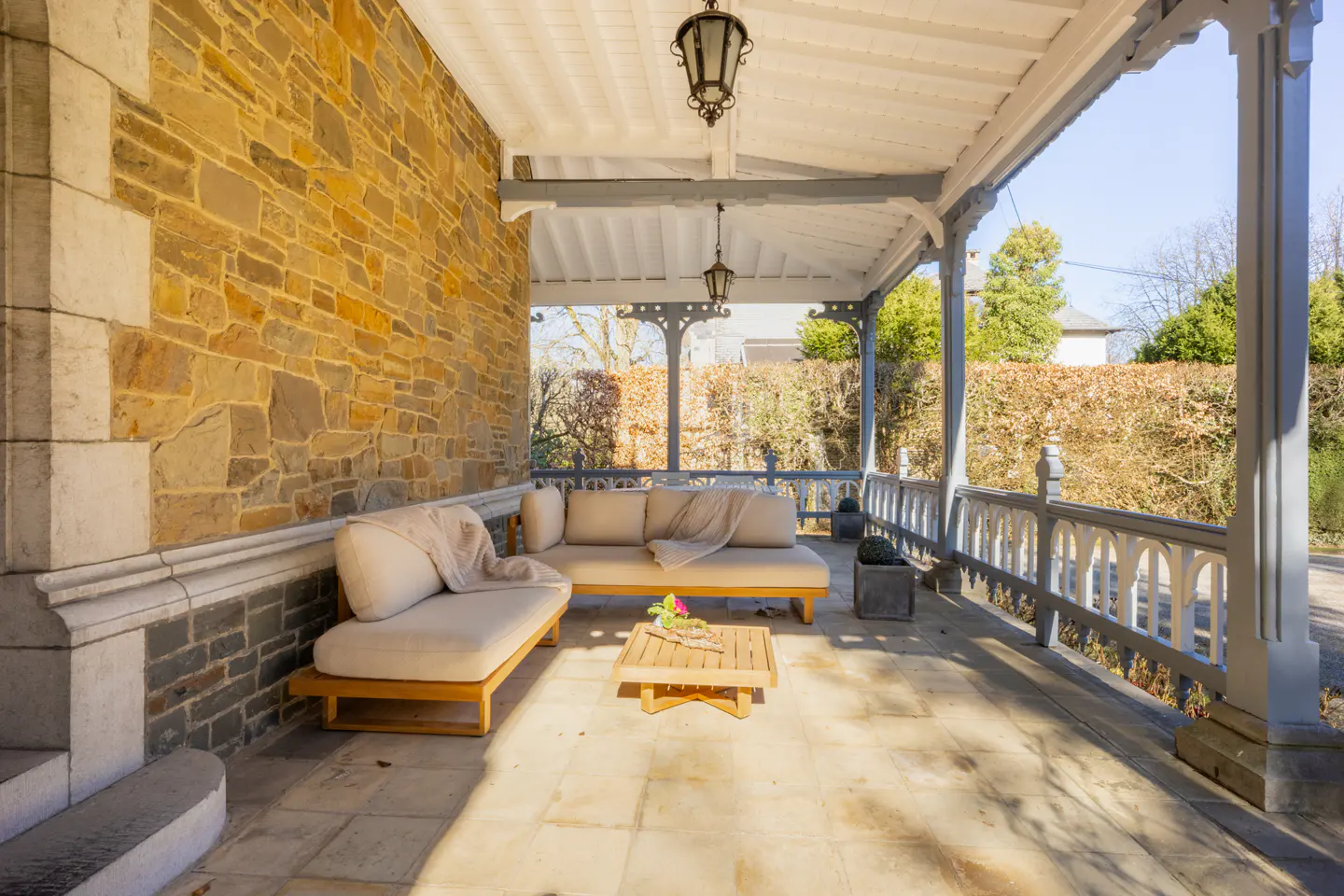 Covered porch with stone wall, gray railings, and outdoor furniture. Beige sofas and a wooden table sit on the stone floor.