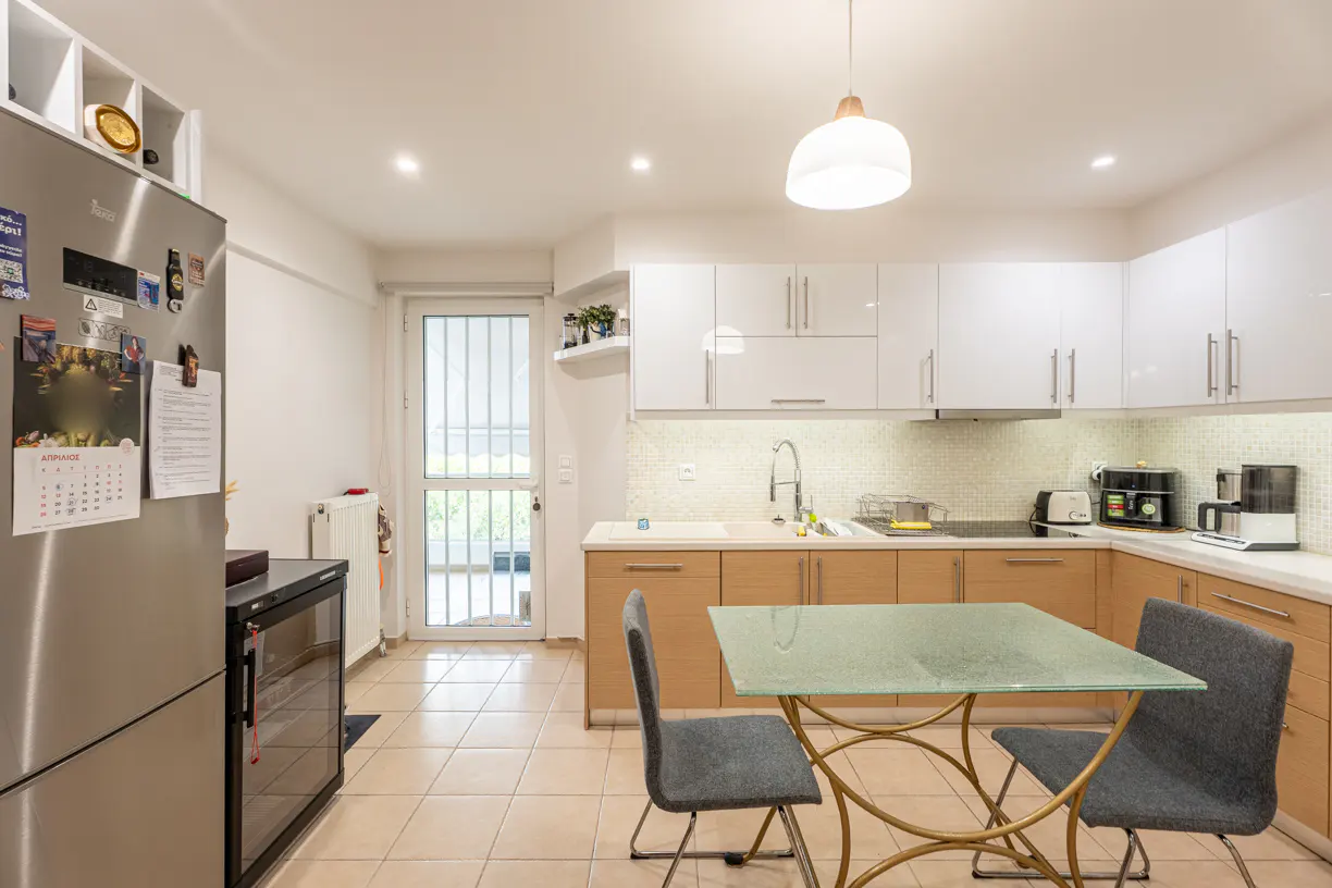 Bright kitchen with white cabinets, light wood lower cabinets, and a glass-topped table with gray chairs. Stainless steel refrigerator on the left.