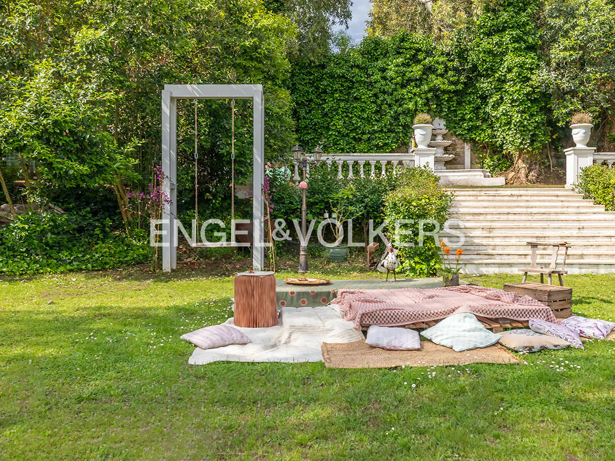 Lush green lawn with a picnic setup: blankets, pillows, and a wooden swing frame. Stairs lead to a balustrade and greenery.