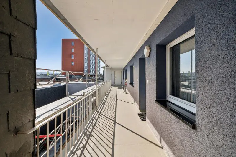 A long, narrow balcony with a white railing and a gray textured wall. Windows line the wall, reflecting the outside view.