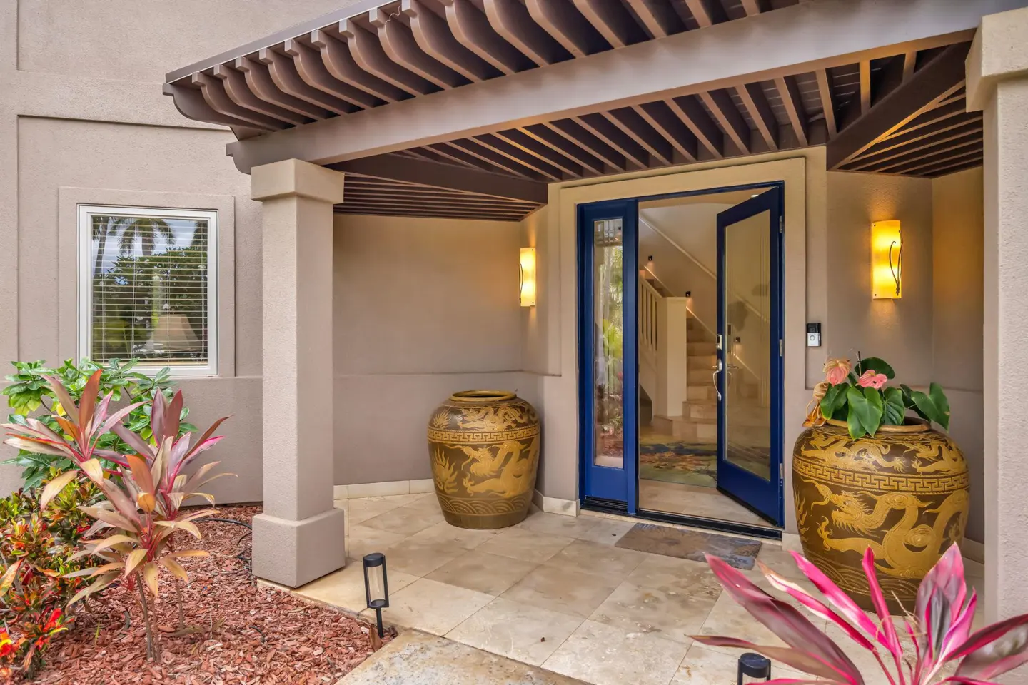 Front entrance with a blue door, pergola, two large decorative gold dragon pots, and tropical plants on a stone porch.