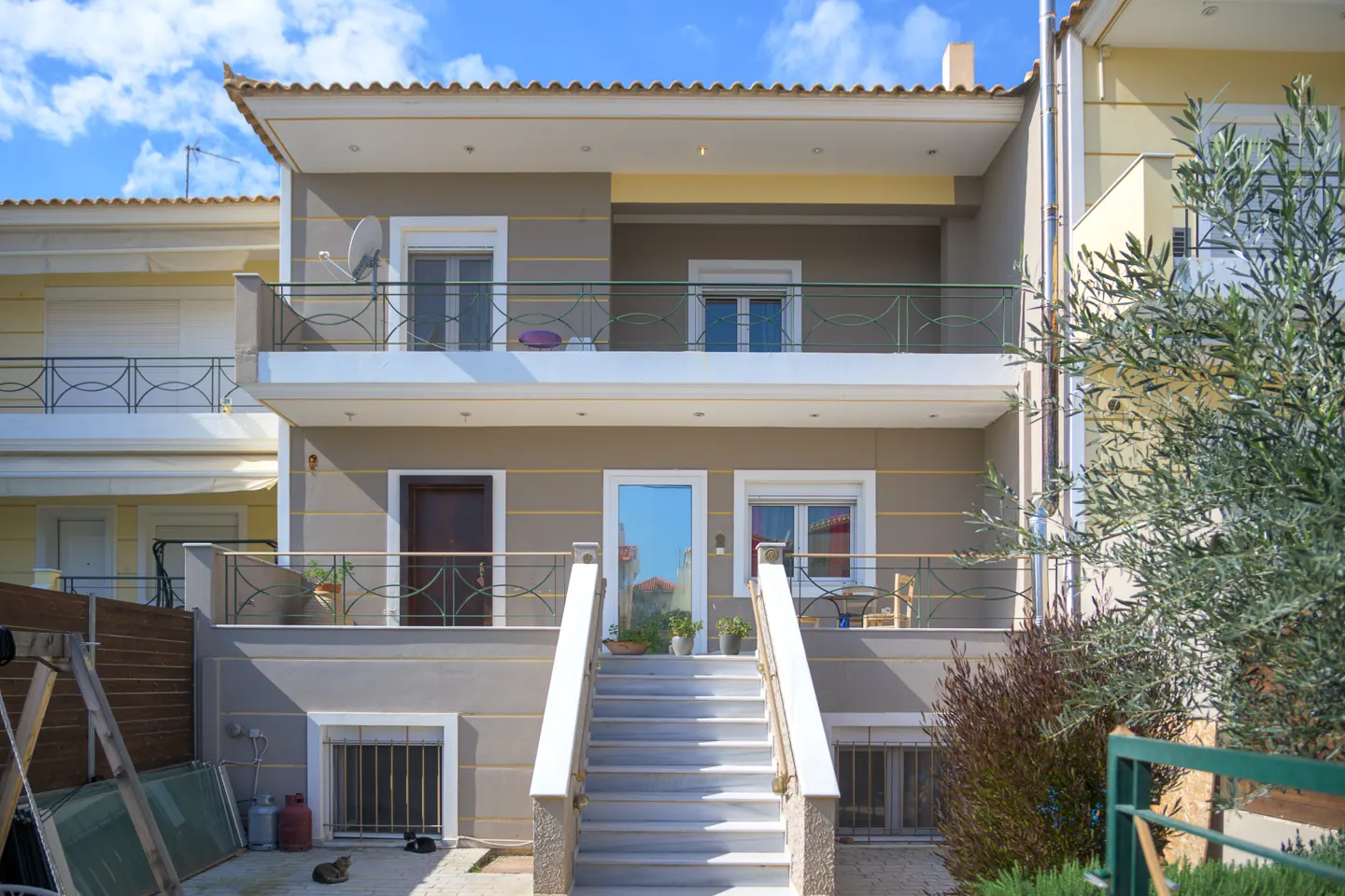 Two-story gray house with white trim and marble stairs leading to the front door. Balconies with green railings on both floors. Blue sky.