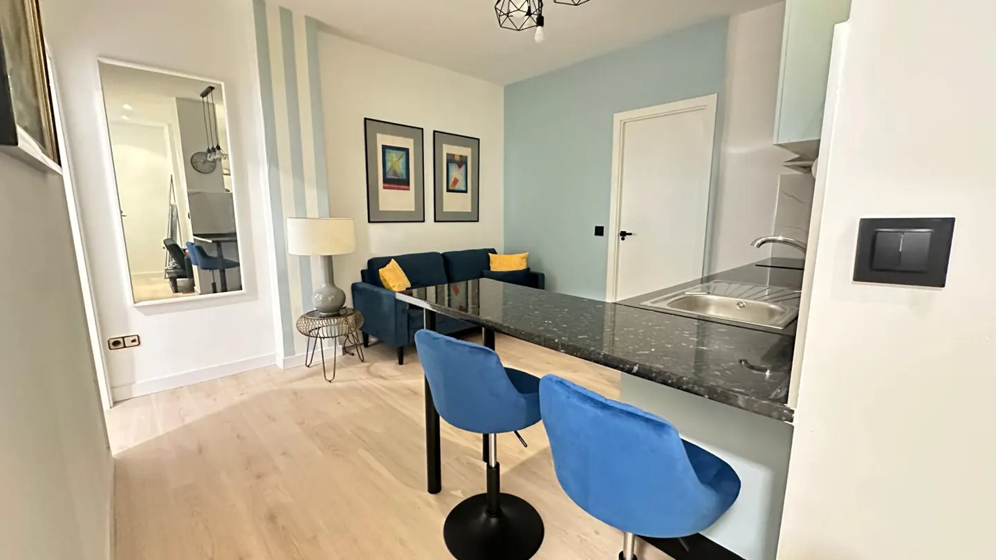 A modern apartment interior with blue velvet chairs at a black granite counter, a blue sofa, and a full-length mirror.