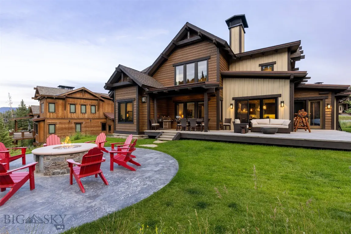 Backyard view of a two-story brown and beige house with a stone fire pit and red Adirondack chairs on a concrete patio.
