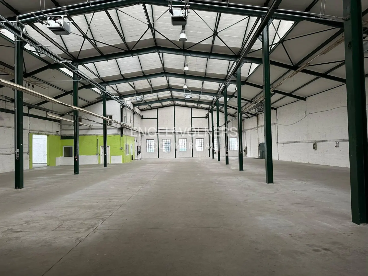 Spacious, empty industrial warehouse with high ceilings, concrete floors, white walls, and green accents.