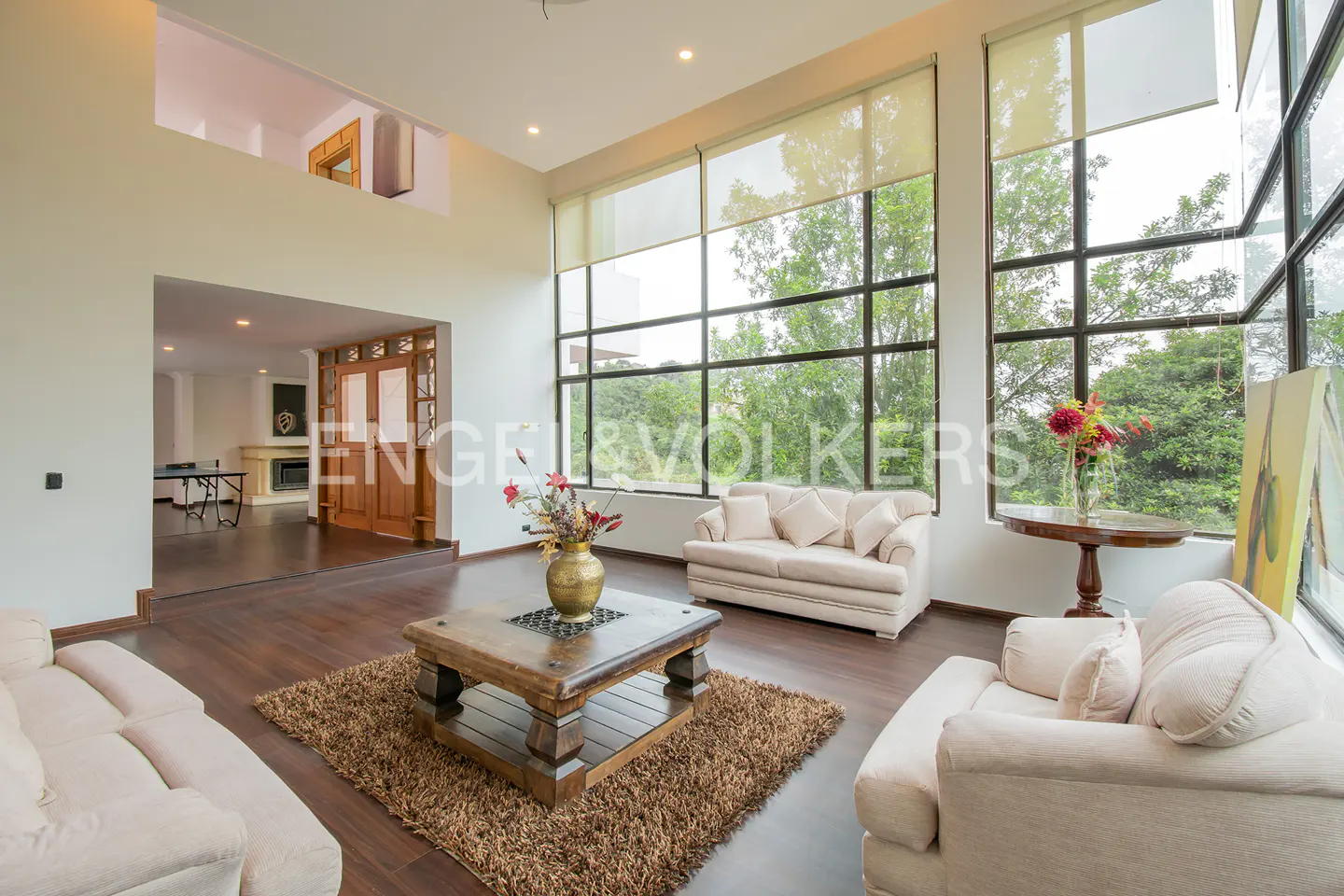 A bright living room with large windows, cream sofas, a wooden coffee table on a brown rug, and a view of lush greenery.
