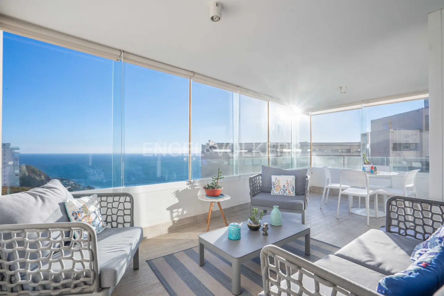 Bright sunroom with ocean view. Gray wicker furniture, glass walls, and a striped rug create a relaxing space.
