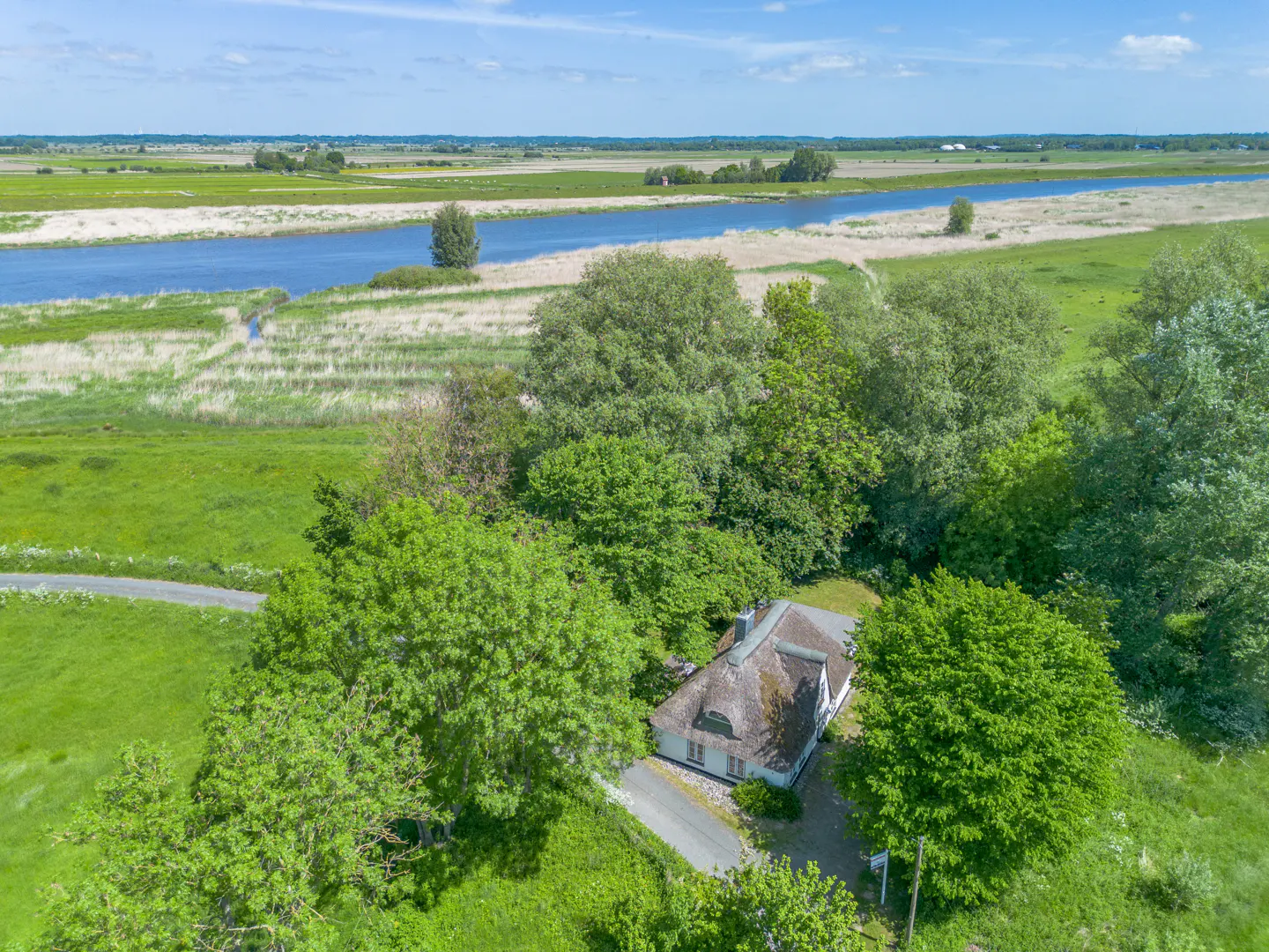 Aerial view of a white house with a thatched roof, surrounded by green trees and a river in the background.