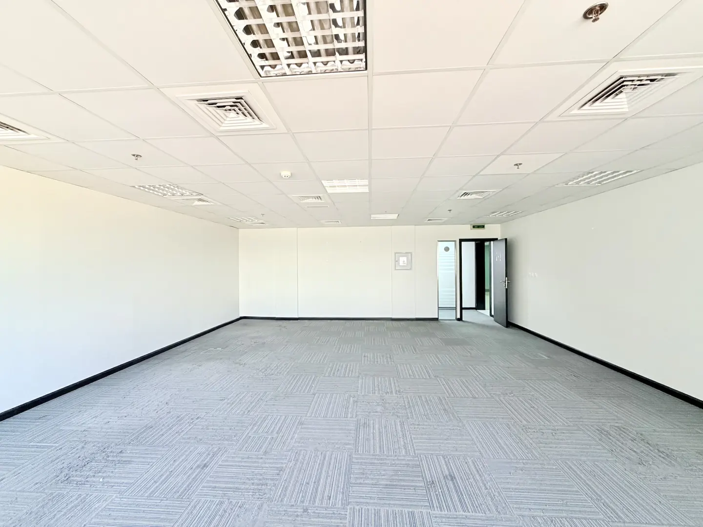 Empty office space with white walls, grey carpet tiles, a drop ceiling with recessed lighting, and an open doorway.