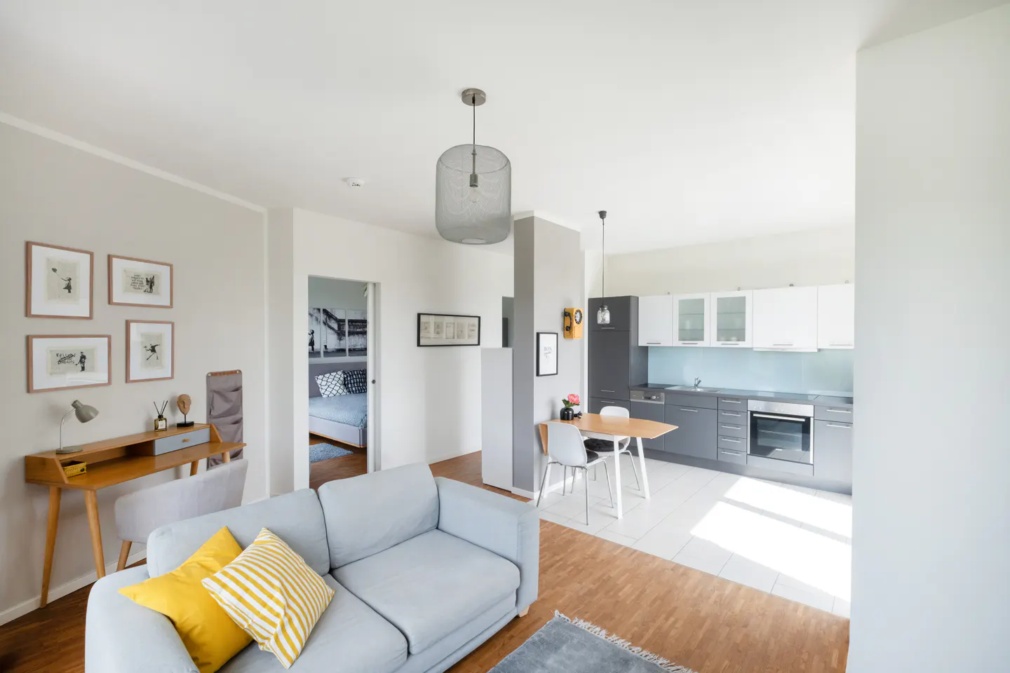 Bright, modern apartment interior with a gray sofa, yellow pillows, desk, and open kitchen with gray cabinets and a wooden dining table.