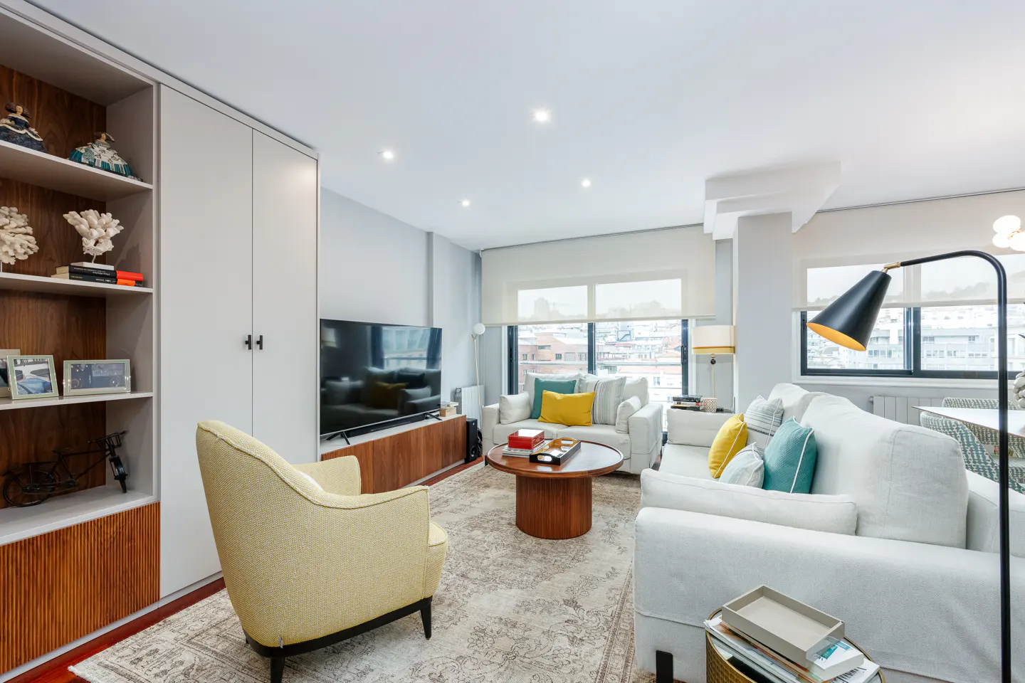Bright living room with white sofas, yellow armchair, and wood accents. Large windows offer city views. Bookshelf and TV add to the cozy space.