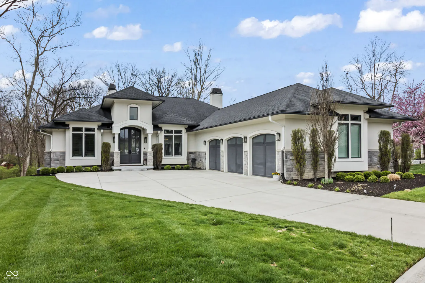 A modern, single-story home with a white exterior, black roof, and three-car garage. A concrete driveway leads to the house, surrounded by green grass and trees.