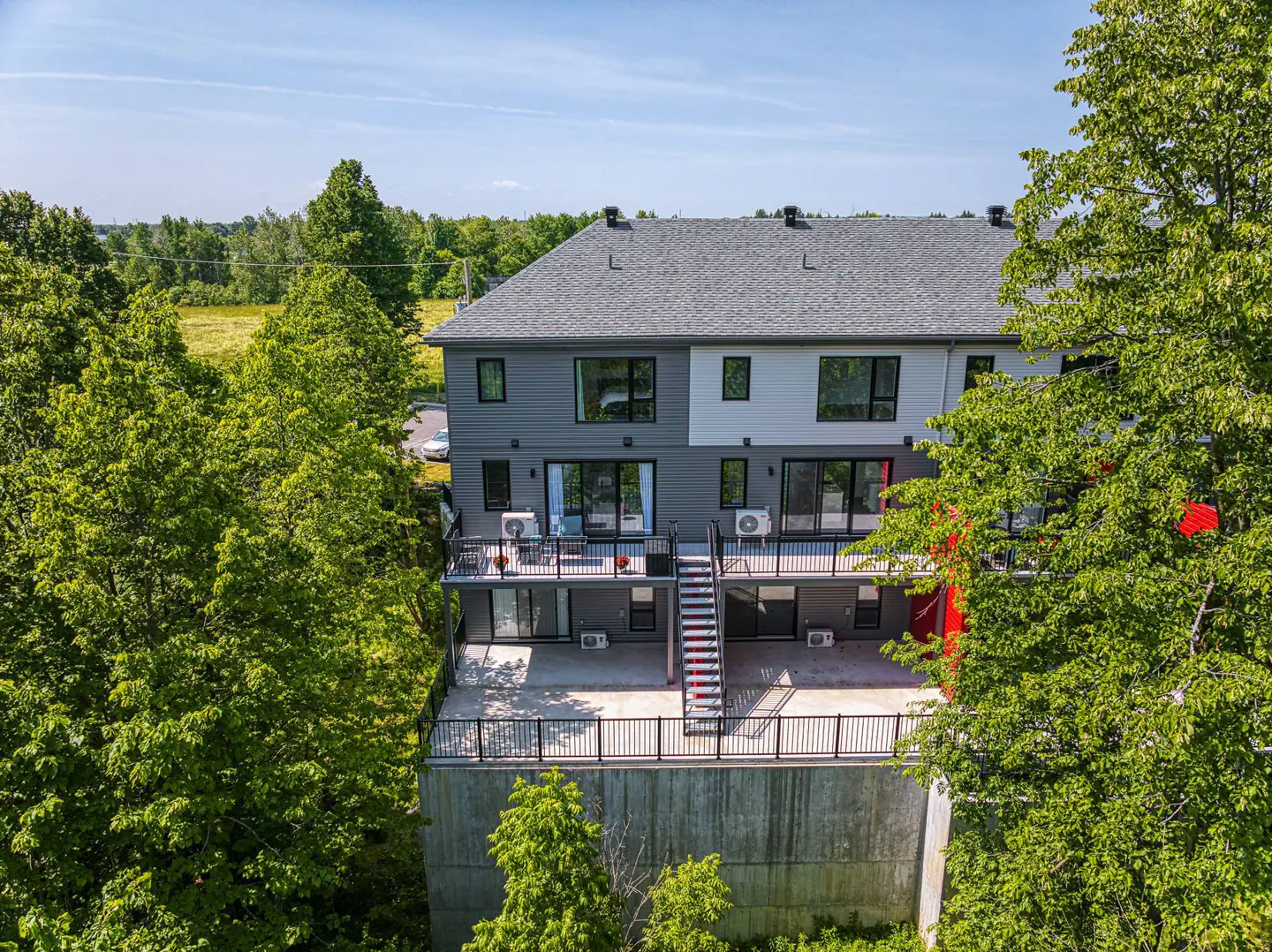 Aerial view of a modern two-story townhouse with gray and white siding, surrounded by lush green trees. It features balconies and a gray roof.