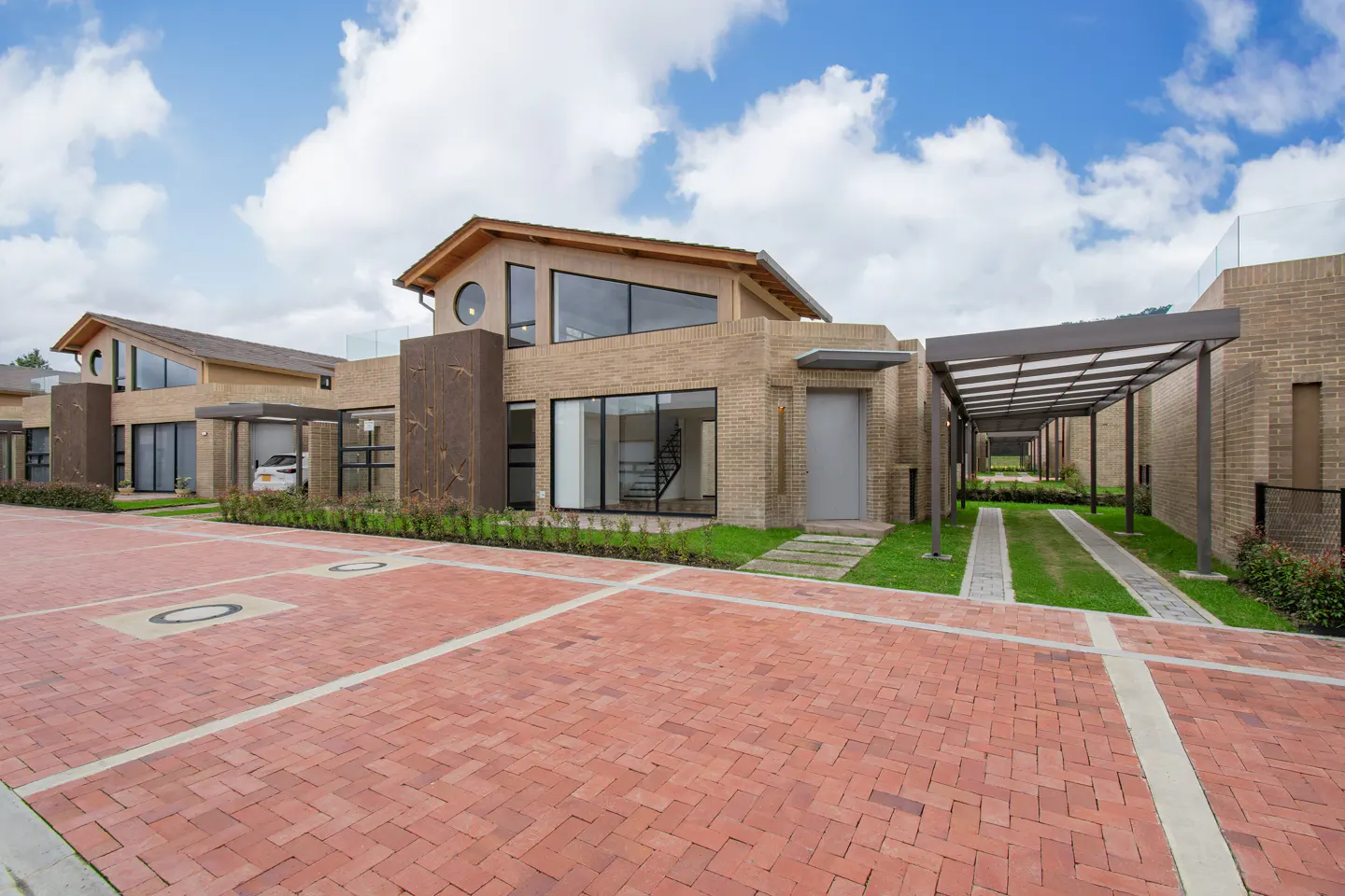 Modern beige brick homes line a red brick street under a blue, cloudy sky. Carports and green grass strips add to the upscale neighborhood feel.