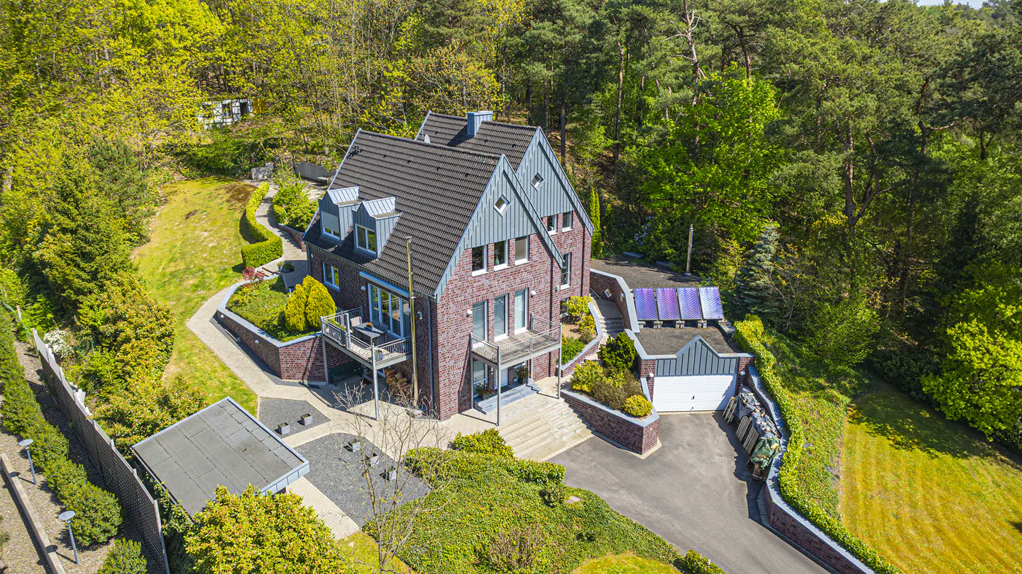 Aerial view of a red brick house with a gray roof, surrounded by lush green trees and manicured landscaping.