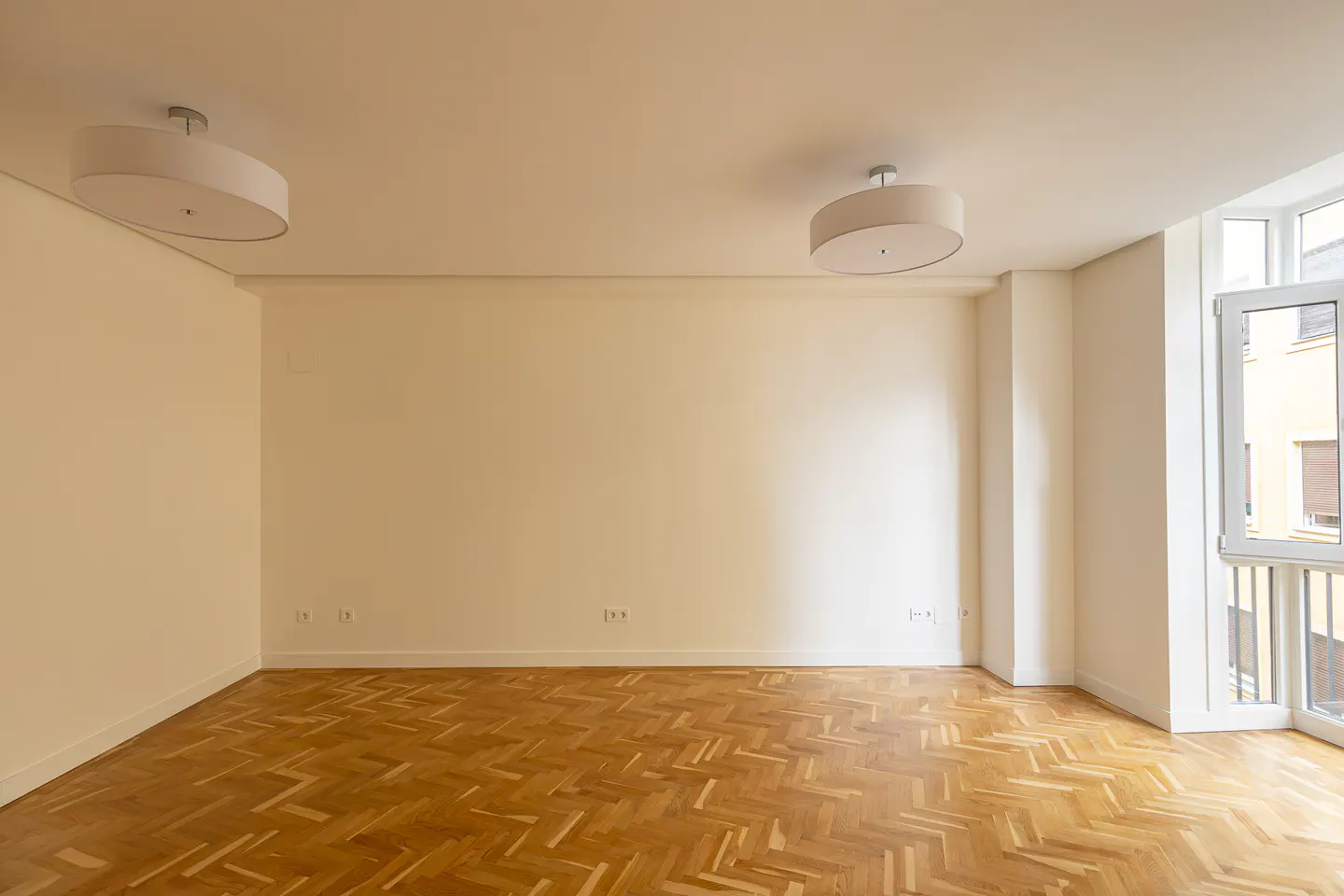 Bright, empty room with herringbone wood floors, white walls, and two drum-shaped ceiling lights. A window with a balcony is on the right.