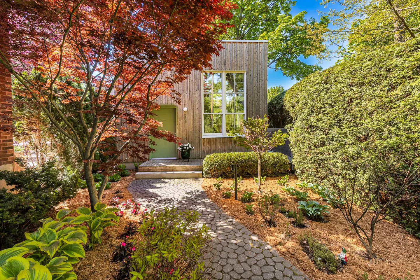 Exterior of a modern wood home with a green door, large window, and stone walkway surrounded by lush landscaping.