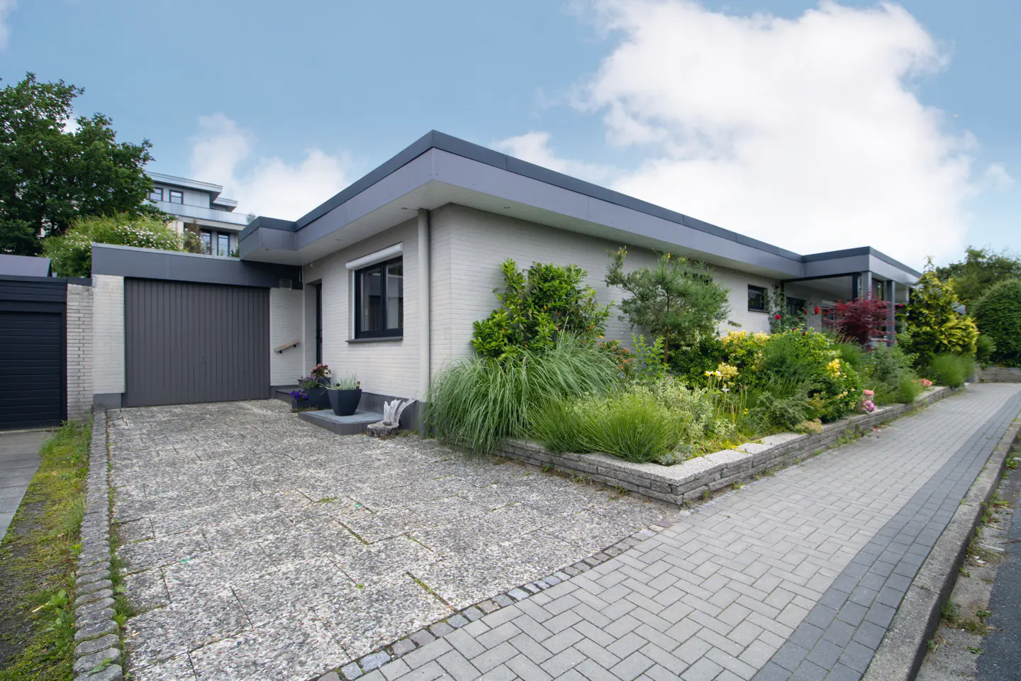 Modern single-story house with a grey brick exterior, flat roof, attached garage, and a landscaped front garden.
