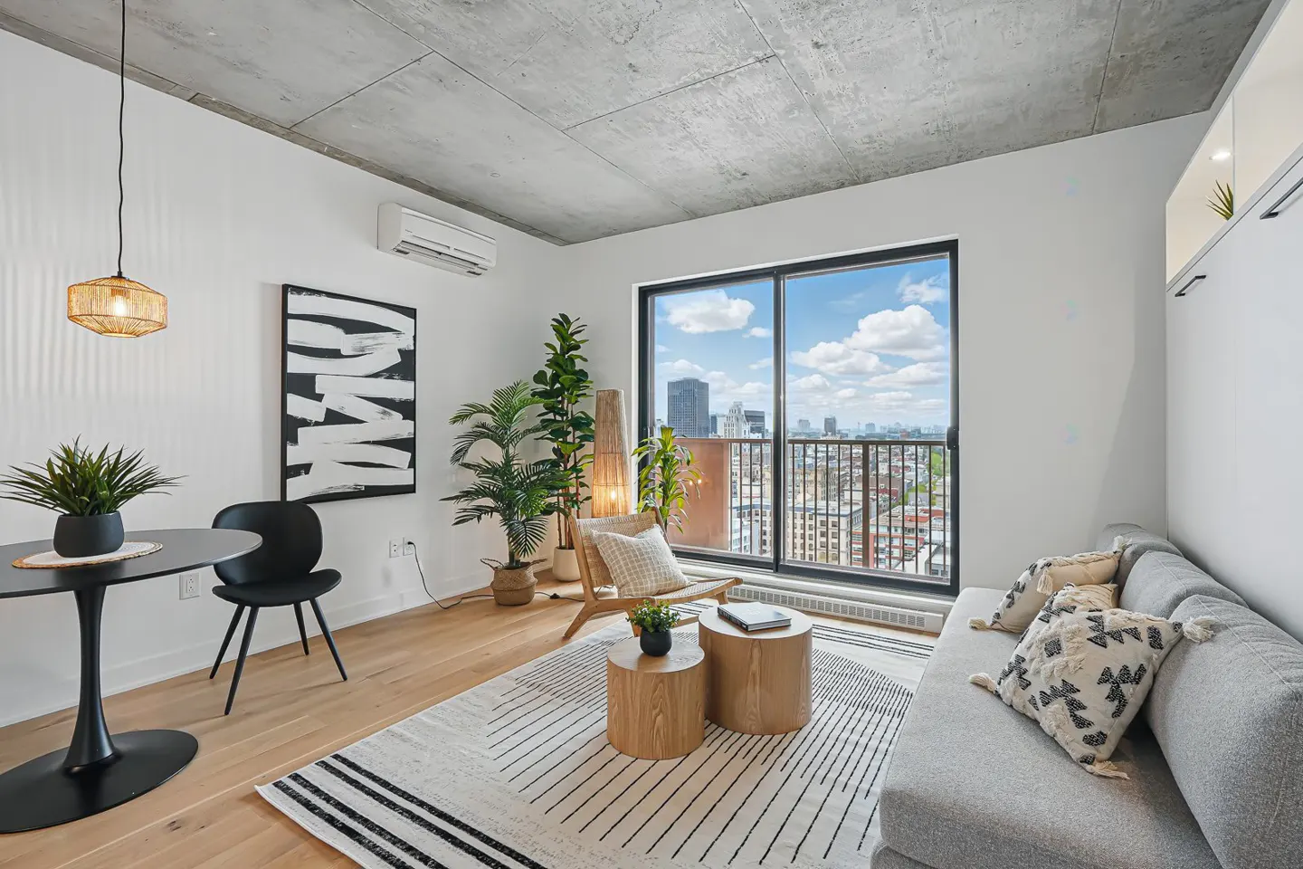 Modern living room with concrete ceiling, grey sofa, round wooden coffee tables, and a balcony view of the city skyline.