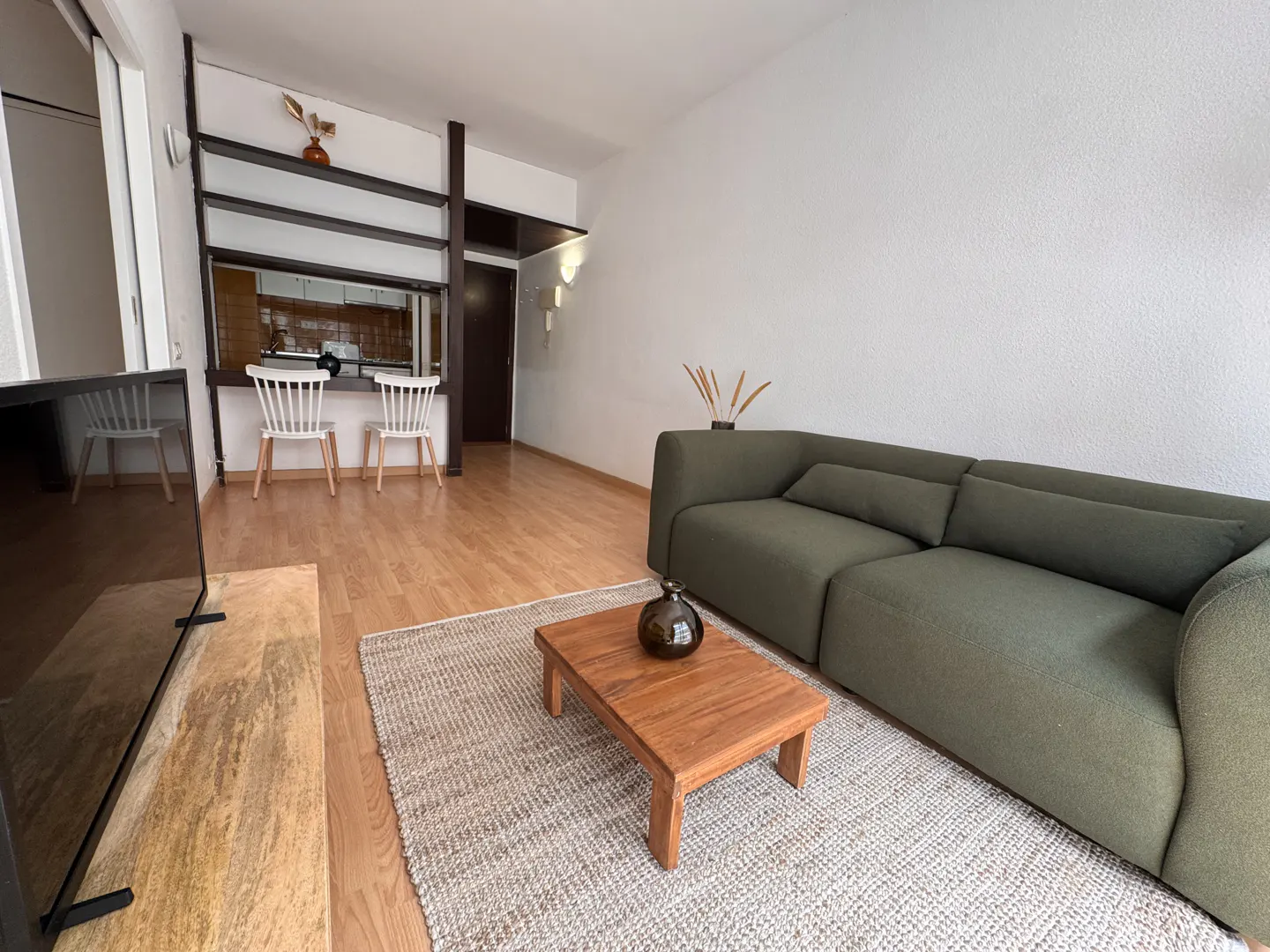 Living room with a green sofa, wooden coffee table, and a beige rug on a light wood floor. Two white chairs are visible through a window to another room.