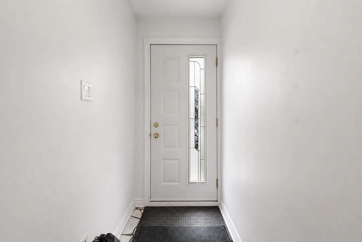 A white front door with a glass panel is centered in a white-walled entryway. A black mat lies in front of the door.