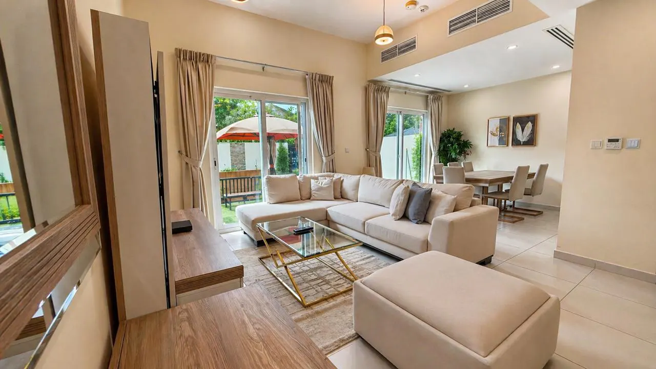 A bright living room with a beige sectional sofa, glass coffee table, and a view of a green backyard through sliding glass doors.