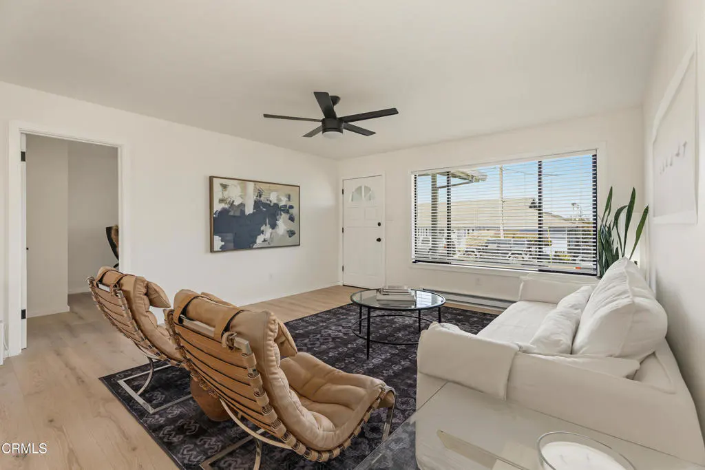 Bright living room with white walls, wood floors, and a black ceiling fan. Two tan chairs face a white sofa and round glass table on a patterned rug.
