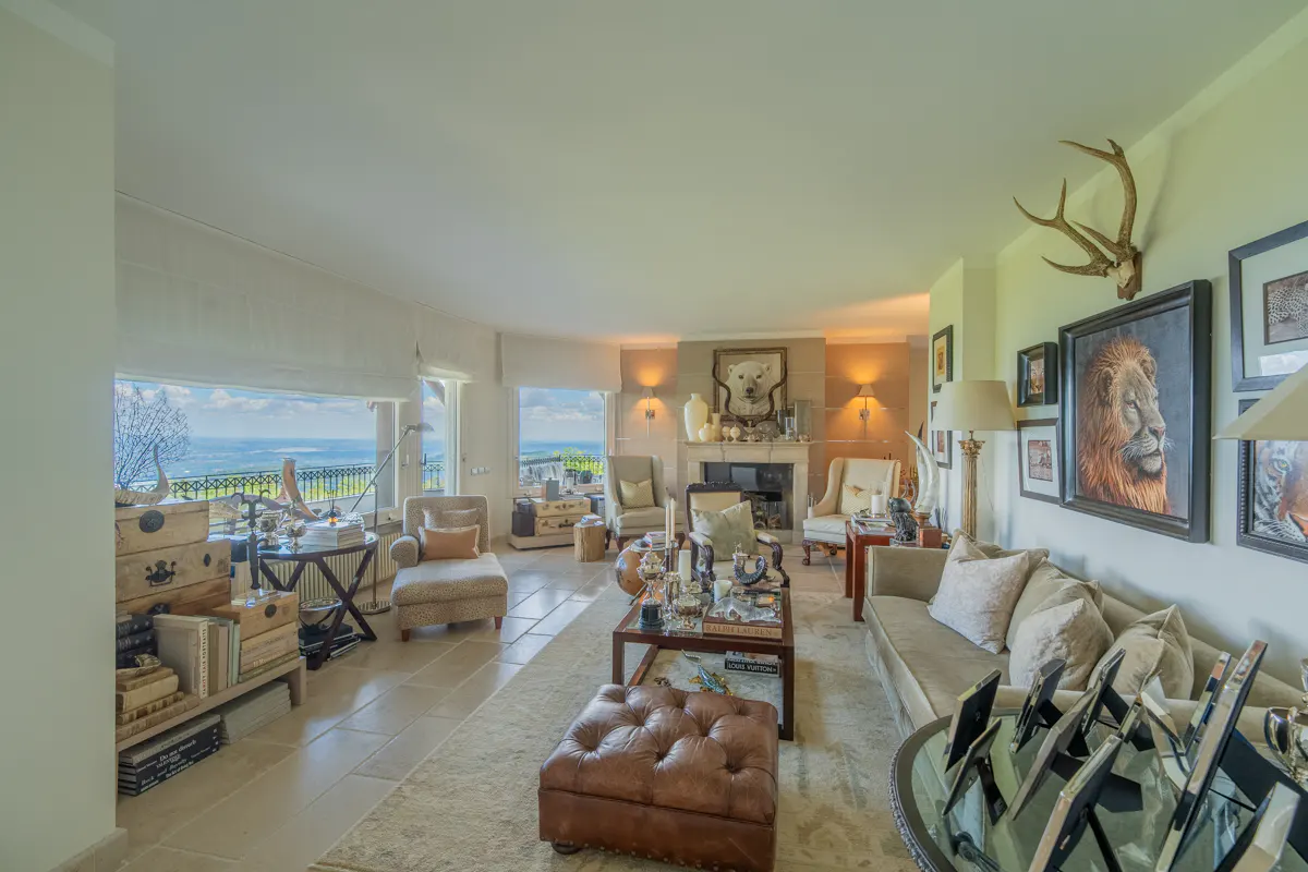 Bright living room with beige sofa, chairs, and ottoman. Large windows offer a scenic view. Walls are decorated with art, including a lion portrait and antlers.