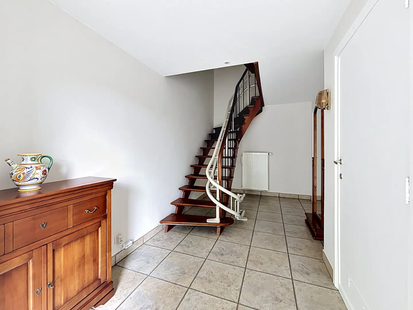 Entryway with tiled floor, wooden cabinet with decorative pitcher, and wooden staircase featuring a white stairlift.