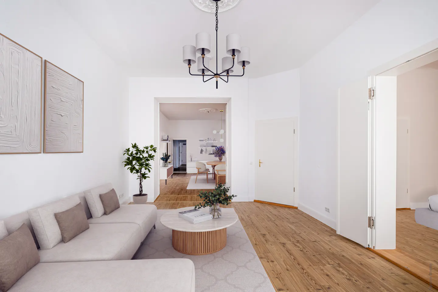 Bright living room with white walls, wood floors, and a beige sectional sofa. A round coffee table sits on a patterned rug, and a modern chandelier hangs from the ceiling.