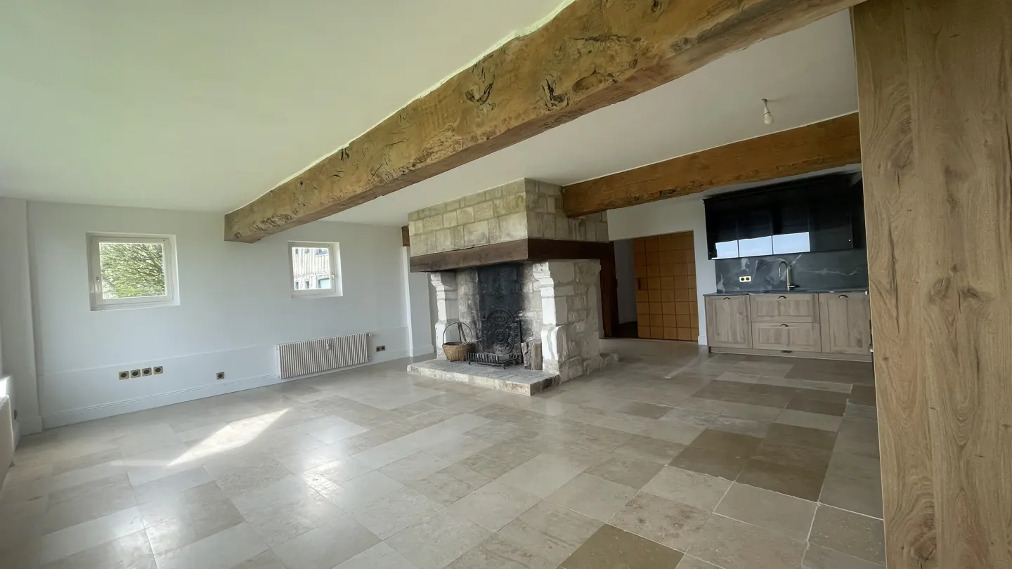Bright, empty living room with stone floors, white walls, and exposed wood beams. A stone fireplace is centered, with a modern kitchen in the background.