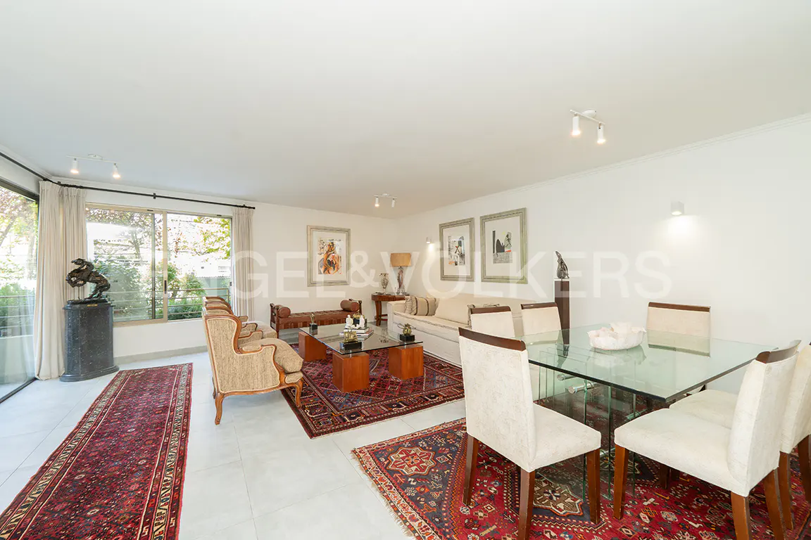 Bright living room with white walls, red rugs, glass table, and cream-colored furniture. Large windows offer a view of greenery.
