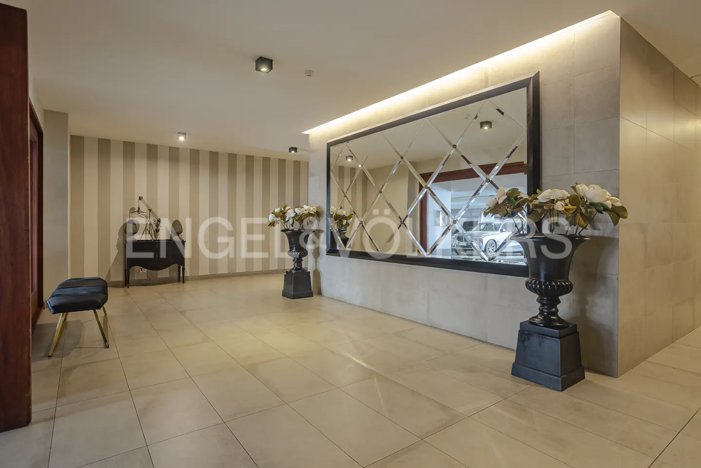 Elegant lobby with beige tile floor, striped wallpaper, and a large mirror. Black urns with white flowers flank the mirror.